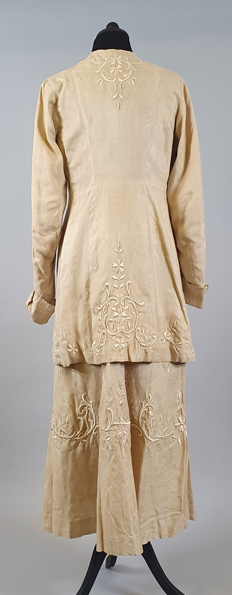 Naturfarget drakt av lin, fôret med silke, bestående av jakke og skjørt. Det følger også med et underskjørt av silke. Jakken er buet over brystet og nederst foran og er innsvinget i livet, og har lange ermer med mansjetter, og har engelsk broderi både på forsiden og på ryggen. Skjørtet har samme type broderi som jakken,