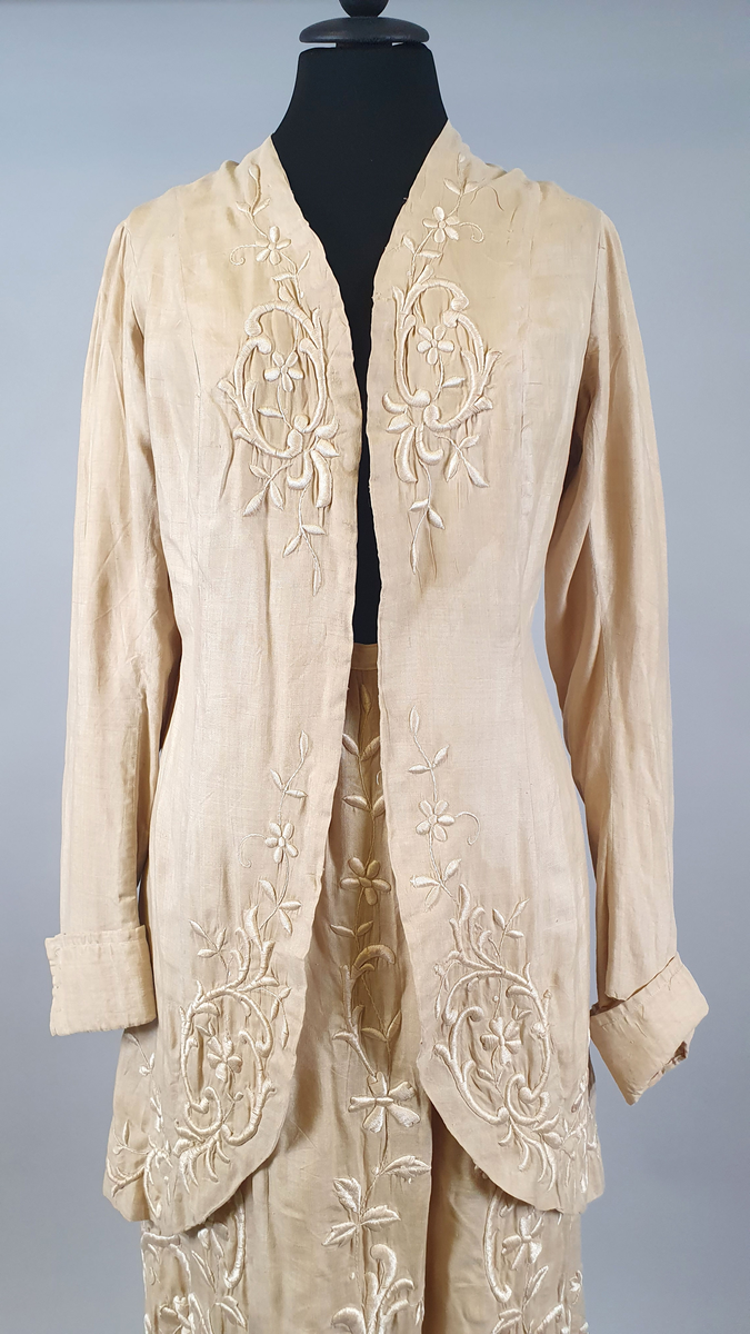 Naturfarget drakt av lin, fôret med silke, bestående av jakke og skjørt. Det følger også med et underskjørt av silke. Jakken er buet over brystet og nederst foran og er innsvinget i livet, og har lange ermer med mansjetter, og har engelsk broderi både på forsiden og på ryggen. Skjørtet har samme type broderi som jakken,