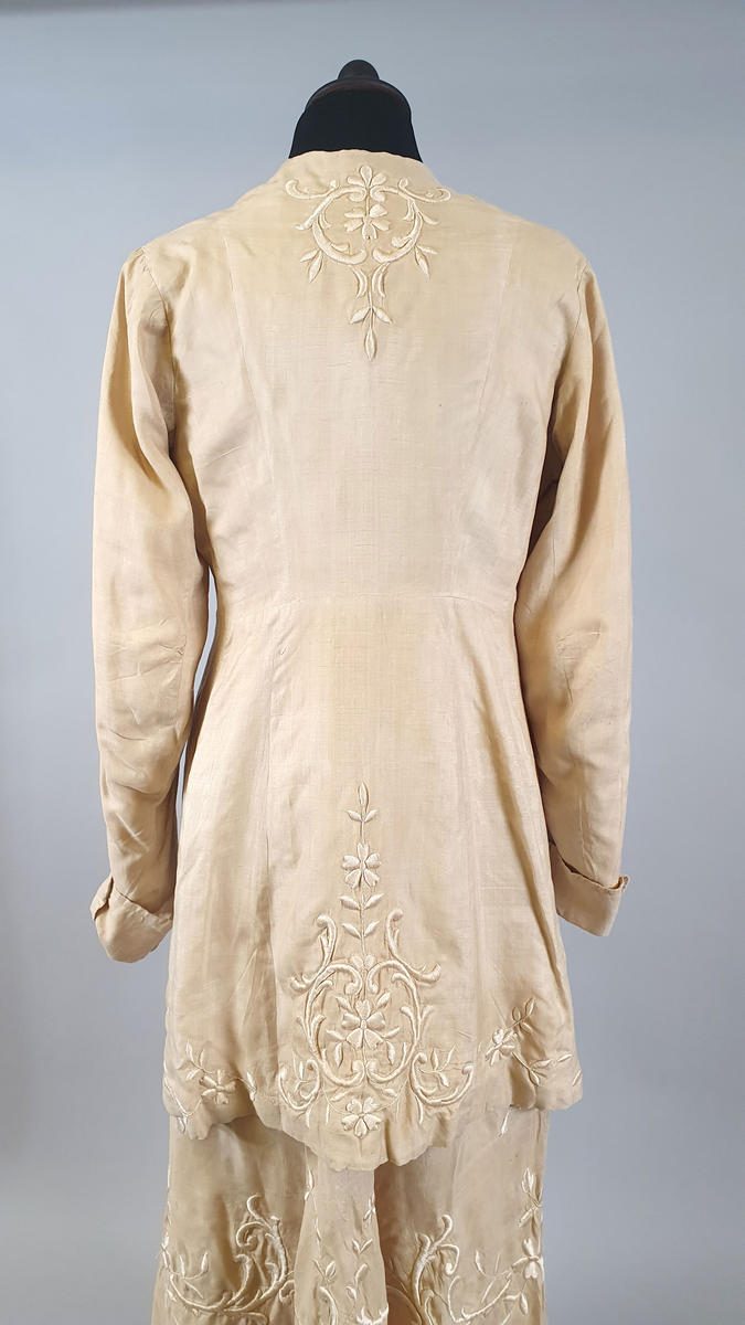 Naturfarget drakt av lin, fôret med silke, bestående av jakke og skjørt. Det følger også med et underskjørt av silke. Jakken er buet over brystet og nederst foran og er innsvinget i livet, og har lange ermer med mansjetter, og har engelsk broderi både på forsiden og på ryggen. Skjørtet har samme type broderi som jakken,