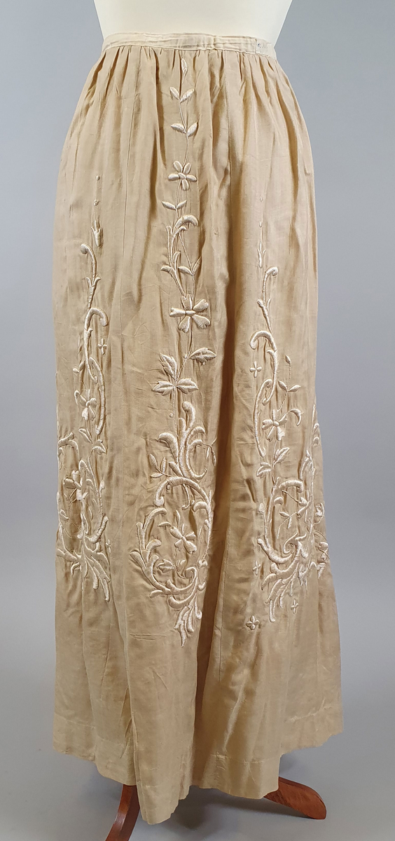 Naturfarget drakt av lin, fôret med silke, bestående av jakke og skjørt. Det følger også med et underskjørt av silke. Jakken er buet over brystet og nederst foran og er innsvinget i livet, og har lange ermer med mansjetter, og har engelsk broderi både på forsiden og på ryggen. Skjørtet har samme type broderi som jakken,