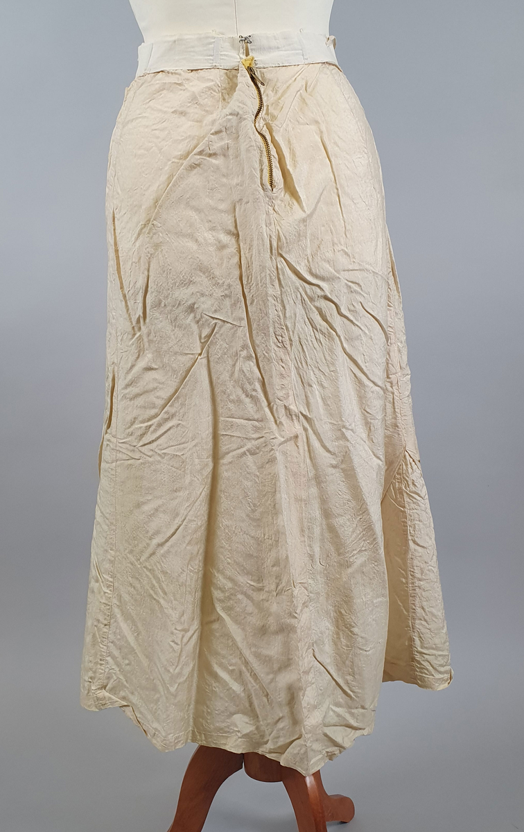 Naturfarget drakt av lin, fôret med silke, bestående av jakke og skjørt. Det følger også med et underskjørt av silke. Jakken er buet over brystet og nederst foran og er innsvinget i livet, og har lange ermer med mansjetter, og har engelsk broderi både på forsiden og på ryggen. Skjørtet har samme type broderi som jakken,