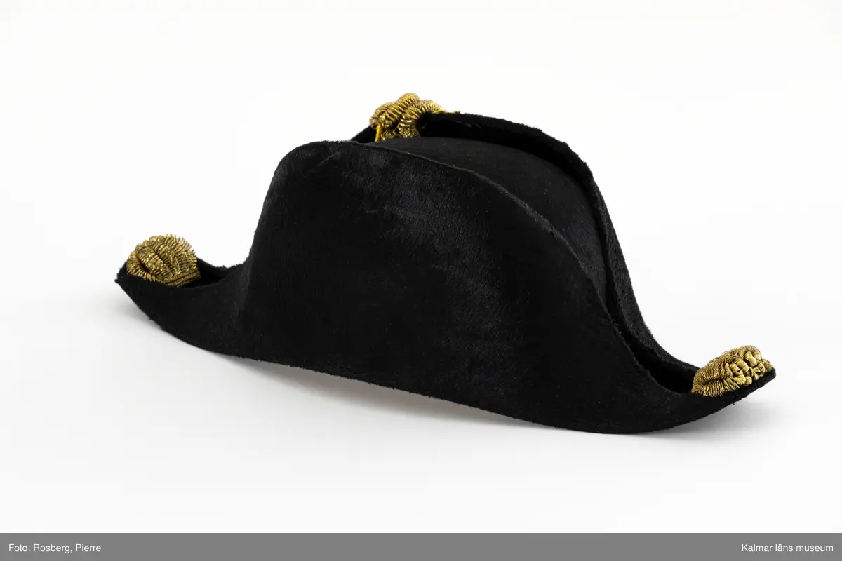 KLM 16548:3:1. Hatt. Bicorne. För revisionssekreterare. Trekantshatt av svart fälb. Dekorerad på höger brätte med gul och blå kokard med guldträns och knapp av gulmetall, knapp med tre kronoroch lagerkrans. I båda ändarna knutformade dekorationer av gulmetallsnodd. Svettrem av ljust skinn. Kullen fodrad med vitt satinvävt siden.  Hakrem av svartfärgat skinn med spänne av gulmetall. Liten vit knapp samt tamp av svart band fastsydda i kullens foder, för att fästa upp hakremmen med. Tryckt svart tillverkaremblem i fodret, under knäpptampen, med text; H.K.H. KRONPRINSENS HOFLEVERANTÖR A.F. BODECKER FABRIKSMÄRKE 24 FREDSGATAN 24 STOCKHOLM.
Till hatten har förvarats i hattask KLM 16548:3:2.