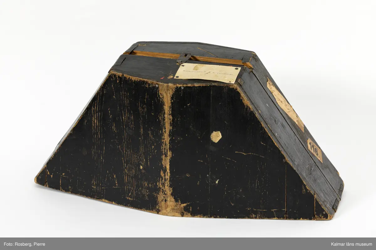 KLM 16895:3:2. Hattask. Fodral till Bicorne för kronofogde. Ask av svartmålat trä. Trekantig form, formad efter hatten. Kan öppnas helt, sammanfogad i botten med två st. gångjärn av vitmetall. Omålad insida. Stängs på ovansidan med någon typ av metallspänne, en av delarna saknas. Visitkort med texten KRONOFOGDEN S. J. A. CEDERBLAD. fastnubbat på askens ovansida. På askens ena sida är tre etiketter fastklistrade. En av dem är oläslig. En har handskriven text Högsby och den tredje etiketten har tryckt text Fr. Lönneberga. 116. N.O.J.
Bicorne KLM 16895:3:1 har förvarats i hattasken.