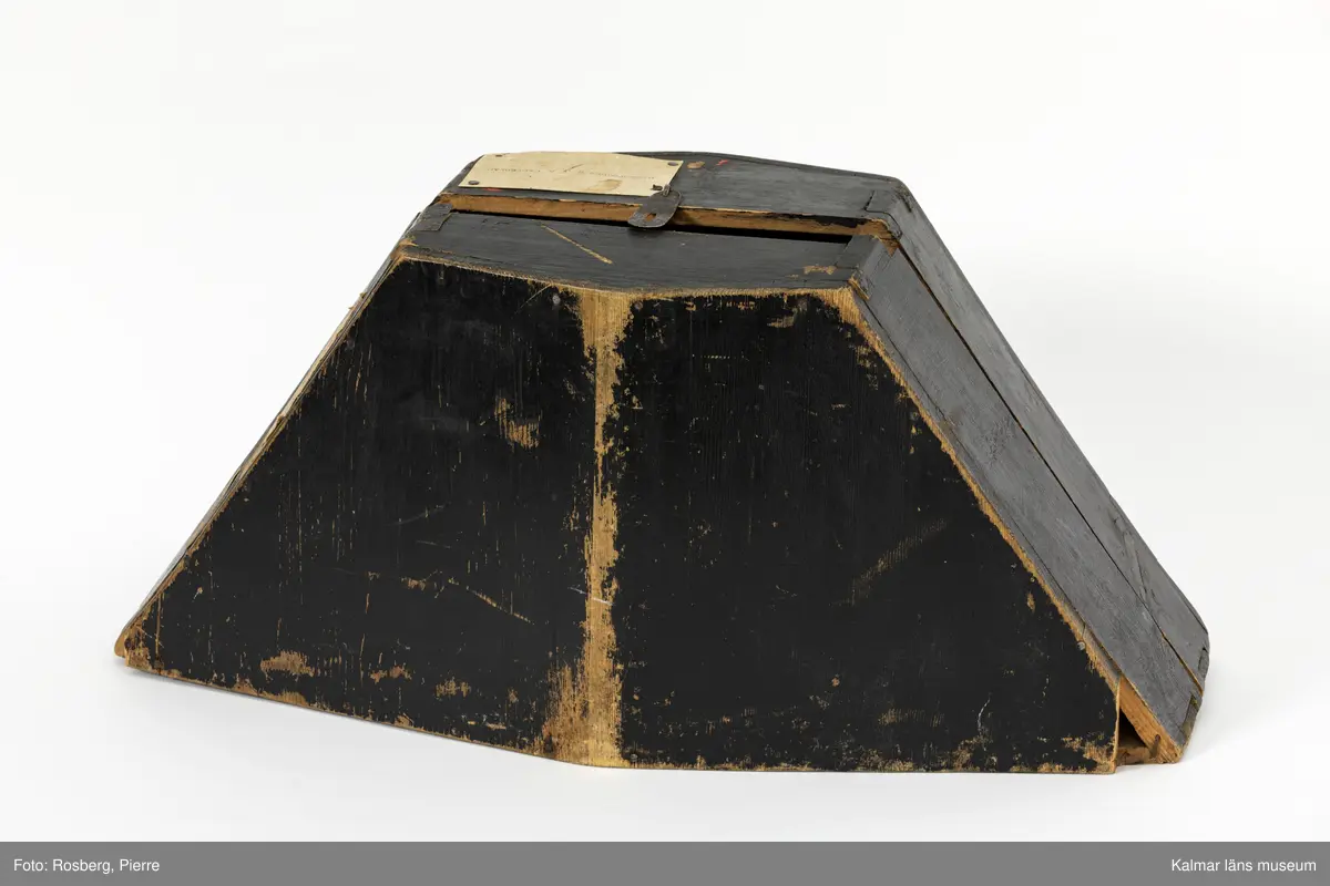 KLM 16895:3:2. Hattask. Fodral till Bicorne för kronofogde. Ask av svartmålat trä. Trekantig form, formad efter hatten. Kan öppnas helt, sammanfogad i botten med två st. gångjärn av vitmetall. Omålad insida. Stängs på ovansidan med någon typ av metallspänne, en av delarna saknas. Visitkort med texten KRONOFOGDEN S. J. A. CEDERBLAD. fastnubbat på askens ovansida. På askens ena sida är tre etiketter fastklistrade. En av dem är oläslig. En har handskriven text Högsby och den tredje etiketten har tryckt text Fr. Lönneberga. 116. N.O.J.
Bicorne KLM 16895:3:1 har förvarats i hattasken.