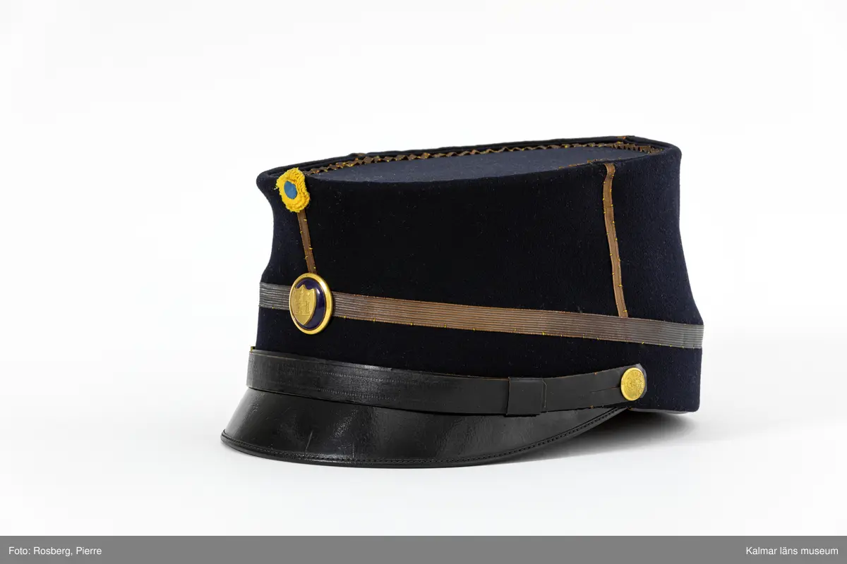 KLM 21690:3. Mössa. Käppi. För poliskommisarie. Av militär modell. Mössa av mörkblått kläde. Rak i sidorna. Platt kulle. Skärm fram av svartlackat läder. Fyra st. enkla stolpar av mörk guldgalon på kullens sidor. Liten gul och blå kokard av siden mitt fram med tre plisserade ringar. Mössknapp under kokarden med Kalmar stads vapen. Mössrem av svart skinn med spänne av gulmetall ovanför skärmen, fästs i mössan med liten kulknapp av gulmetall med Kalmar läns vapen. En rad guldgalon nära mössans nederkant. Svettrem av brunt skinn. Kullen fodrad med blank svart satinvävd bomull. Tryckt text i guld i fodret: MILITÄR EKIPERINGS AKTIEBOLAGET STOCKHOLM.
