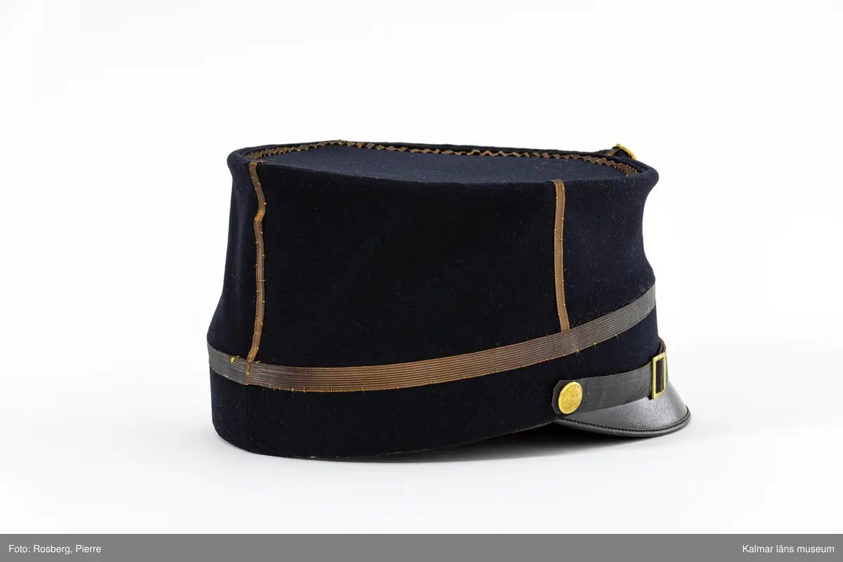 KLM 21690:3. Mössa. Käppi. För poliskommisarie. Av militär modell. Mössa av mörkblått kläde. Rak i sidorna. Platt kulle. Skärm fram av svartlackat läder. Fyra st. enkla stolpar av mörk guldgalon på kullens sidor. Liten gul och blå kokard av siden mitt fram med tre plisserade ringar. Mössknapp under kokarden med Kalmar stads vapen. Mössrem av svart skinn med spänne av gulmetall ovanför skärmen, fästs i mössan med liten kulknapp av gulmetall med Kalmar läns vapen. En rad guldgalon nära mössans nederkant. Svettrem av brunt skinn. Kullen fodrad med blank svart satinvävd bomull. Tryckt text i guld i fodret: MILITÄR EKIPERINGS AKTIEBOLAGET STOCKHOLM.