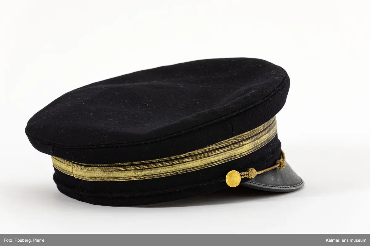 KLM 21693. Mössa. Skärmmössa. För poliskommisarie. Av svart kläde. Cirkelrund platt kulle med något överskjutande sidor ovanför mössans raka kanter. Svart skärm mitt fram. På mössans kant en bred guldgalon med smal ripsvävd svart mittrand. På galonen gul blå kokard med tre st. plisserade ringar. Ovanför kokarden murkrona av gulmetalltråd broderad på mörkblått kläde fastsytt på mössan mitt fram. Mössrem av guldsnodd ovanför skärmen, knäpps i mössan med gulmetallknapp med murkrona och eklövskrans. Svettrem av ljusbrunt skinn. Innanför svettremmen fastsydd pappremsa. Kullen fodrad med svart sativävt tyg med rombformade mönsterstickningar. Vävd svart tillverkaretikett med text i gult; Alb. Palmquist HATT MÖSS & PÄLSAFFÄR KALMAR, fastsydd i fodret.