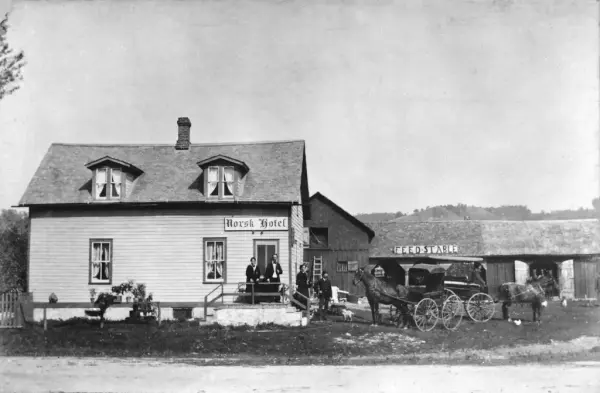 Historisk bilde av en én-etasjes bygning med skilt "Norsk Hotel". Tre mennesker på trappa utenfor inngangen. To hester og kjerrer til høyre for hotellet. I bakgrunn en driftsbygning med  skilt "feed stable" på taket.