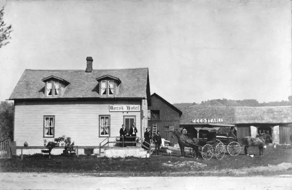 Historisk bilde av en én-etasjes bygning med skilt "Norsk Hotel". Tre mennesker på trappa utenfor inngangen. To hester og kjerrer til høyre for hotellet. I bakgrunn en driftsbygning med  skilt "feed stable" på taket.