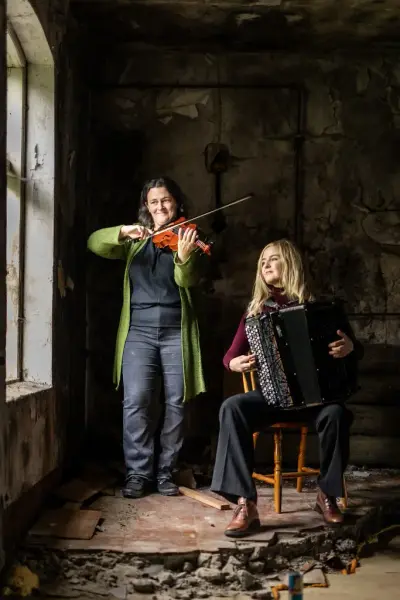 Bilde av folkemusikerne Britt Elise Skram og Kristina Bjørdal. Britt Elise står og holder en fele og Kristina sitter på en trestol og holder ett trekkspill. De er inne i et slitt lokale.