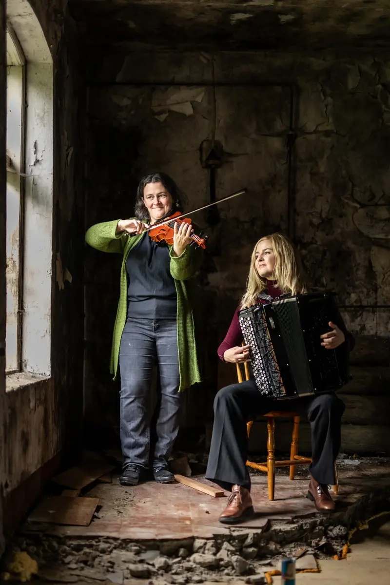 Bilde av folkemusikerne Britt Elise Skram og Kristina Bjørdal. Britt Elise står og holder en fele og Kristina sitter på en trestol og holder ett trekkspill. De er inne i et slitt lokale.