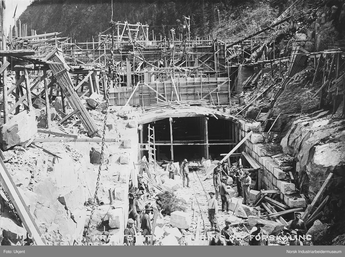 Arbeid med undervannskanal på Vemork Kraftstasjon. Trekonstruksjon rundt på anleggsområdet, klopper for trasnport med trillebår i terrenget. Trakter for tipp av masse fra øverste nivå og ned til arbeiderne i kanalen. Belysning henger i stolper på området.