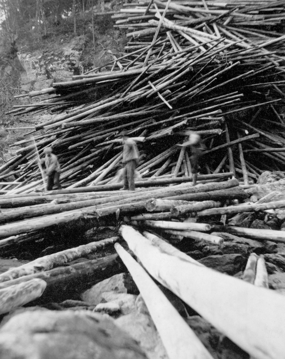 Utislag av tømmer ved Fossum bru i nedre del av Glomma i 1922.  Fotografiet er tatt fra et elveleie der det later til å ha vært svært liten vannføring akkurat da fotografiet ble tatt.  Her lå det et beskjedent antall tømmerstokker på digre steiner, dels på tvers av det som, når flommen kom, ville bli strømretningen.  I den bratte skråningen bakenfor lå det tømmer, som øyensynlig var veltet utfor fra en ovenforliggende veg.  Tømmeret i den bratte skråningen var sammenvaset, og det ville antakelig bli en vanskelig og farefull oppgave for fløterne å få det ut i vassdraget.  Tre mannsskikkelser i forgrunnen var antakelig fløtere som hadde begynt på dette arbeidet før flommen kom med større og striere vannføring. 