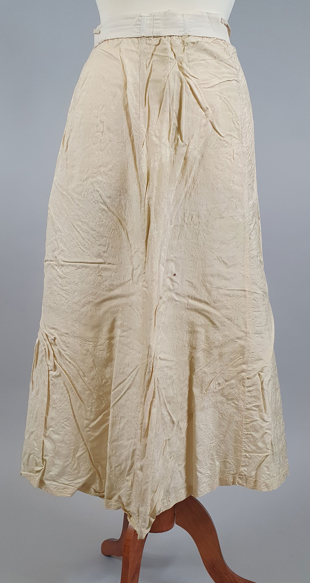 Naturfarget drakt av lin, fôret med silke, bestående av jakke og skjørt. Det følger også med et underskjørt av silke. Jakken er buet over brystet og nederst foran og er innsvinget i livet, og har lange ermer med mansjetter, og har engelsk broderi både på forsiden og på ryggen. Skjørtet har samme type broderi som jakken,