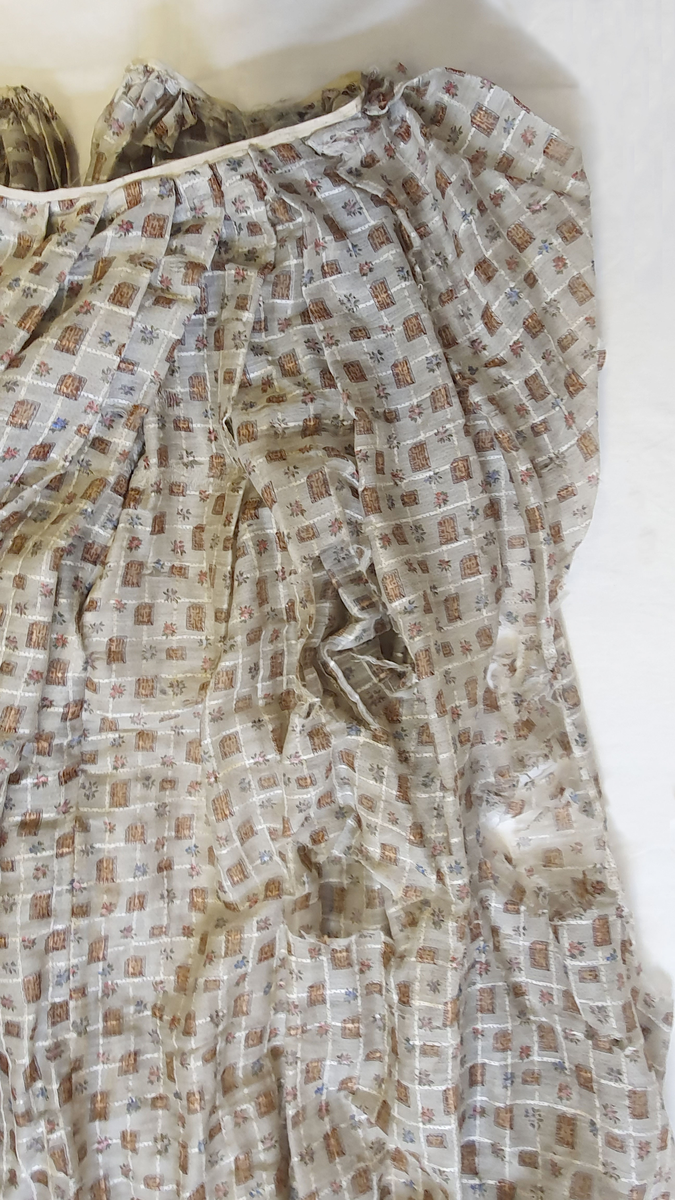 Skjørt av tynn, transparent bomull, med mønster av små kvadrater i hvitt rutenett. Skoningen har tre paralette bånd langs kanten. Stoffet er svært nedbrutt, og det er flere store revner og hull,