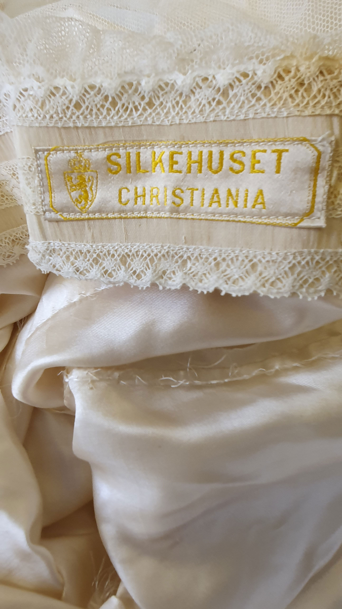 Brudekjole av naturhvit silke i ett stykke. Overdelen består av tyll og silke, der forstykket av silke er løst og må festes på skudrene. Den nedre delen er hoftesid, men sidere på hver side enn midt foran. Belte i livet med metallhemper. Tyll med blonder dekker skudrene og er sydd fast over venste bryst, midt bak er det festet et lodd, og det høyre stykket er løst.