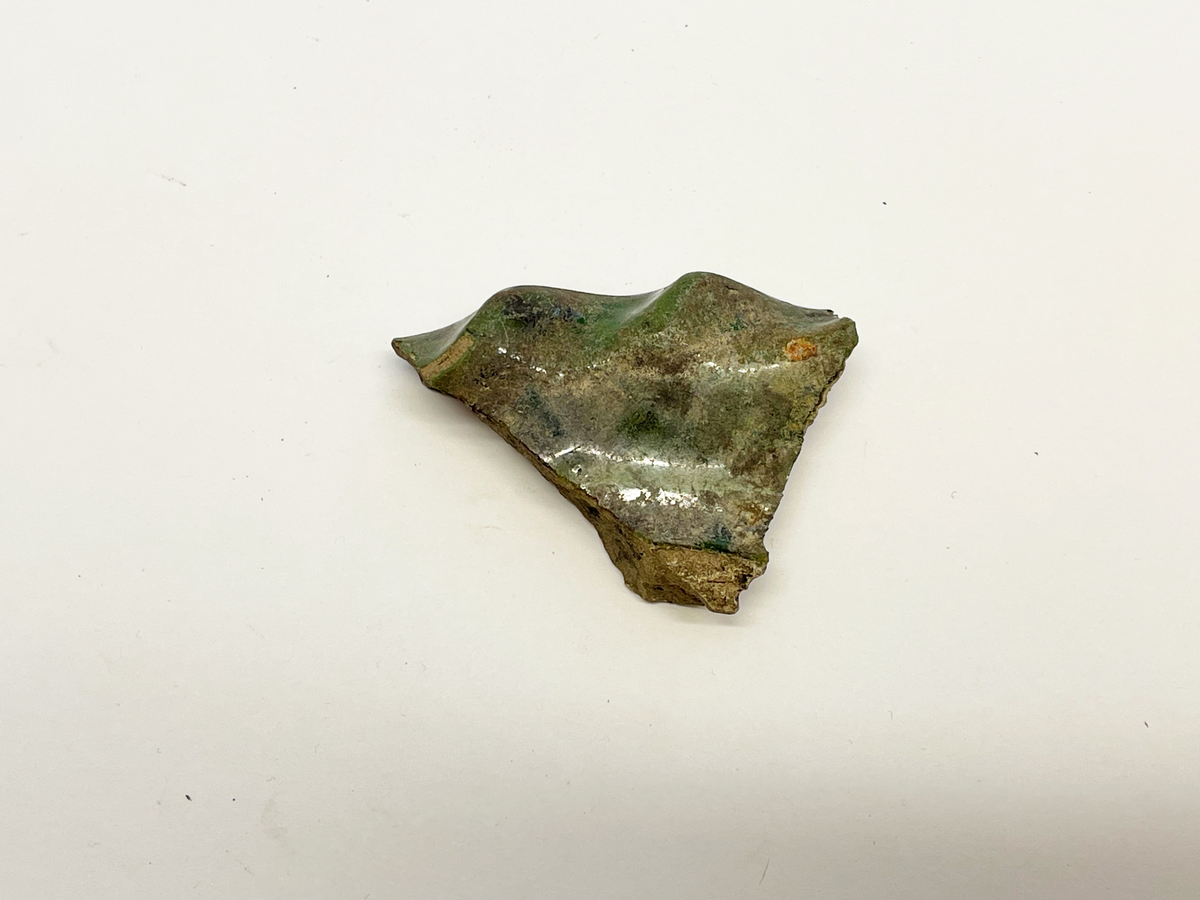 Fotfragment av beige lergods med grön glasyr på ovan- och delar av utsidan. Foten formad som ett 30 mm brett brätte med tummad, vågig kant. Ljusbrännande rödgods eller vitgods? Mycket lik M 48845-26e, 29, 33. 35, 36, 44-49 och 84 i glasyr och gods.
