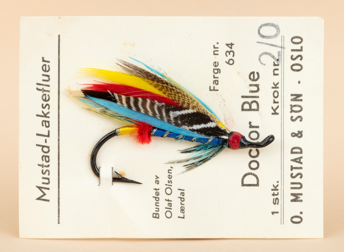 Ubrukt fiskeflue, våtflue, lakseflue, produsert for O. Mustad & Søn av Olaf Olsen. Fiskeflua er bundet på en fabrikkprodusert stålkrok. Den ferdigbundne flua er festet til en rektangulær papplate med påtrykt produktinformasjon (avskrevet i feltet "Påført tekst/merker") og  deretter emballert i en blank plastikkpose (trolig cellofan). Cellofaninnpakningen er skjør og delvis spjæret på ene siden. Det er pklistret en prislapp utenpå innnpakningen med teksten : Bilco Tromsø 12.50

Fiskeflua er bundet på en svart stålkrok som har mothake i den ene enden, et rødfarget hode med oppadbøyd (krokøye) for senefeste i andre enden.  Flua har blåfarget kropp med sølvtinsel (metallglisende striper). I forlengelsen av dette har fluebinderen laget en rød butt og et gulfarget tag. Flua har blågrønt fronthackle med innslag av svart og grått. 
Flua har vinge som er laget av mangefarget fjær: blå, rød, gul, svart med hvit spragling og brun med lysebrun spragling. Vingefjærene strekker seg lengre enn krokbøyen. 

[For informasjon om fiskefluer og fluebinding er blant annet boka "Sportsfiskeleksikon" fra 2005 en kilde. Se også omtale av Olaf Olsen under fanen "Andre opplysninger".]