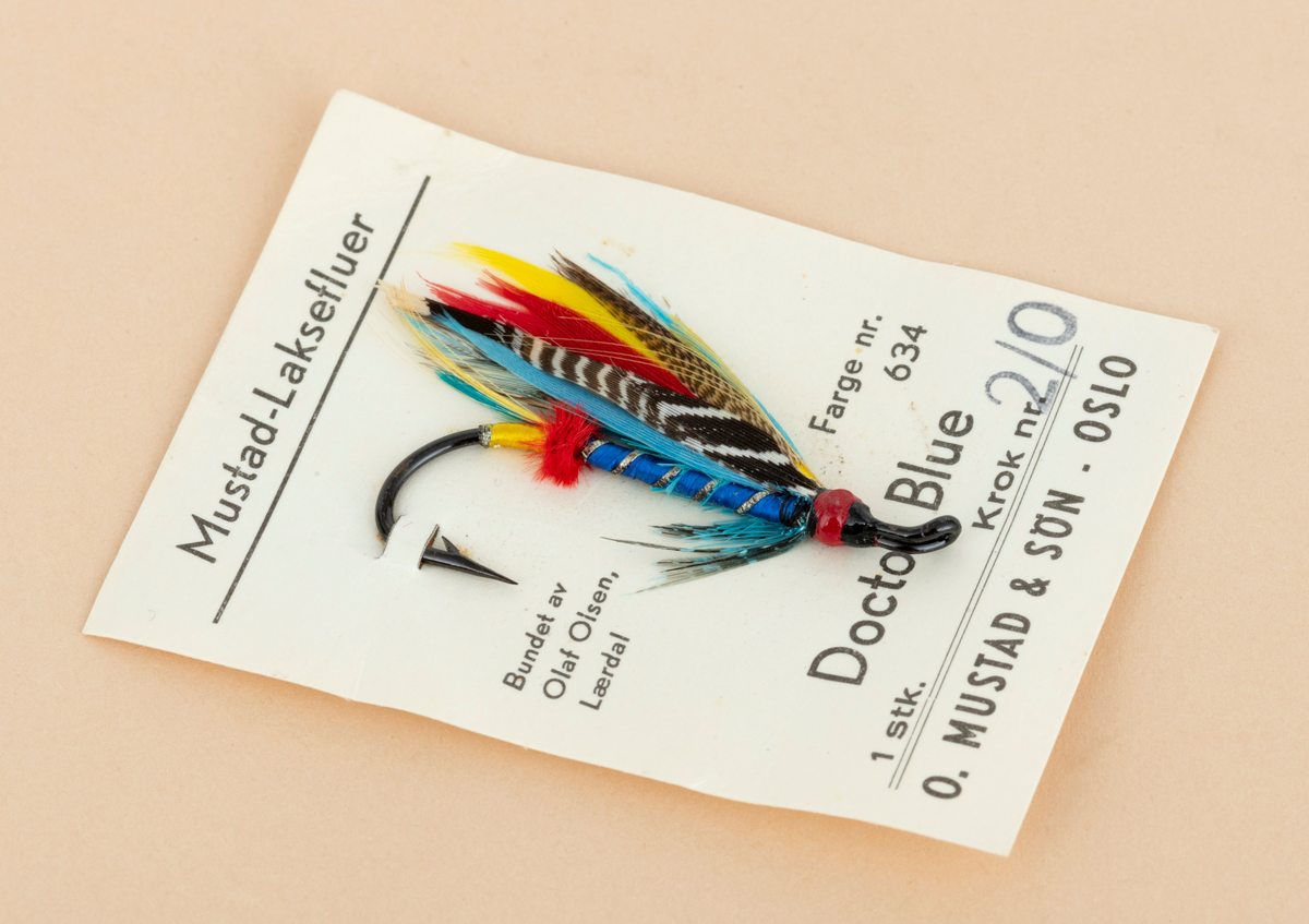 Ubrukt fiskeflue, våtflue, lakseflue, produsert for O. Mustad & Søn av Olaf Olsen. Fiskeflua er bundet på en fabrikkprodusert stålkrok. Den ferdigbundne flua er festet til en rektangulær papplate med påtrykt produktinformasjon (avskrevet i feltet "Påført tekst/merker") og  deretter emballert i en blank plastikkpose (trolig cellofan). Cellofaninnpakningen er skjør og delvis spjæret på ene siden. Det er pklistret en prislapp utenpå innnpakningen med teksten : Bilco Tromsø 12.50

Fiskeflua er bundet på en svart stålkrok som har mothake i den ene enden, et rødfarget hode med oppadbøyd (krokøye) for senefeste i andre enden.  Flua har blåfarget kropp med sølvtinsel (metallglisende striper). I forlengelsen av dette har fluebinderen laget en rød butt og et gulfarget tag. Flua har blågrønt fronthackle med innslag av svart og grått. 
Flua har vinge som er laget av mangefarget fjær: blå, rød, gul, svart med hvit spragling og brun med lysebrun spragling. Vingefjærene strekker seg lengre enn krokbøyen. 

[For informasjon om fiskefluer og fluebinding er blant annet boka "Sportsfiskeleksikon" fra 2005 en kilde. Se også omtale av Olaf Olsen under fanen "Andre opplysninger".]