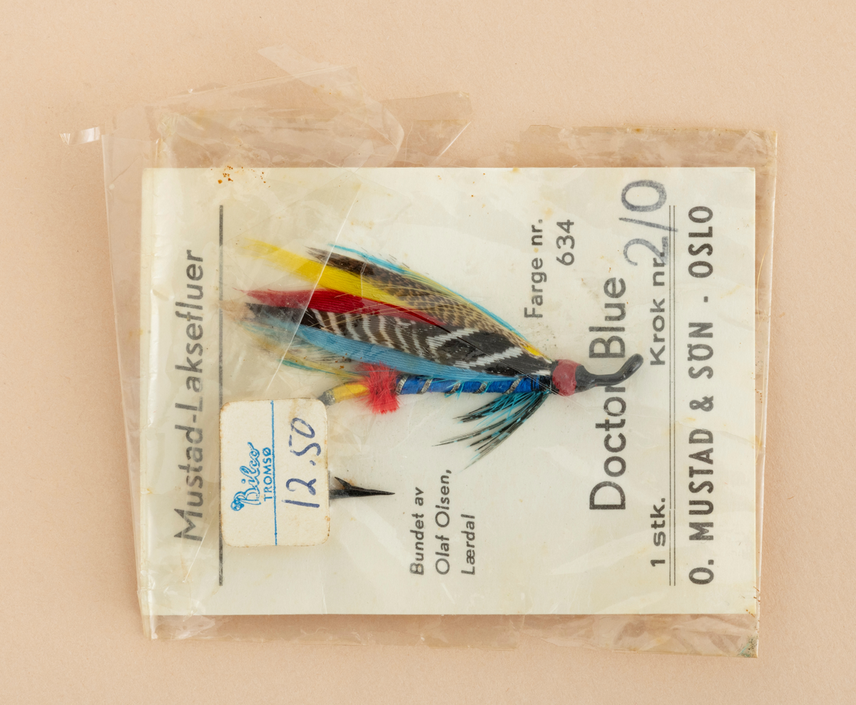 Ubrukt fiskeflue, våtflue, lakseflue, produsert for O. Mustad & Søn av Olaf Olsen. Fiskeflua er bundet på en fabrikkprodusert stålkrok. Den ferdigbundne flua er festet til en rektangulær papplate med påtrykt produktinformasjon (avskrevet i feltet "Påført tekst/merker") og  deretter emballert i en blank plastikkpose (trolig cellofan). Cellofaninnpakningen er skjør og delvis spjæret på ene siden. Det er pklistret en prislapp utenpå innnpakningen med teksten : Bilco Tromsø 12.50

Fiskeflua er bundet på en svart stålkrok som har mothake i den ene enden, et rødfarget hode med oppadbøyd (krokøye) for senefeste i andre enden.  Flua har blåfarget kropp med sølvtinsel (metallglisende striper). I forlengelsen av dette har fluebinderen laget en rød butt og et gulfarget tag. Flua har blågrønt fronthackle med innslag av svart og grått. 
Flua har vinge som er laget av mangefarget fjær: blå, rød, gul, svart med hvit spragling og brun med lysebrun spragling. Vingefjærene strekker seg lengre enn krokbøyen. 

[For informasjon om fiskefluer og fluebinding er blant annet boka "Sportsfiskeleksikon" fra 2005 en kilde. Se også omtale av Olaf Olsen under fanen "Andre opplysninger".]