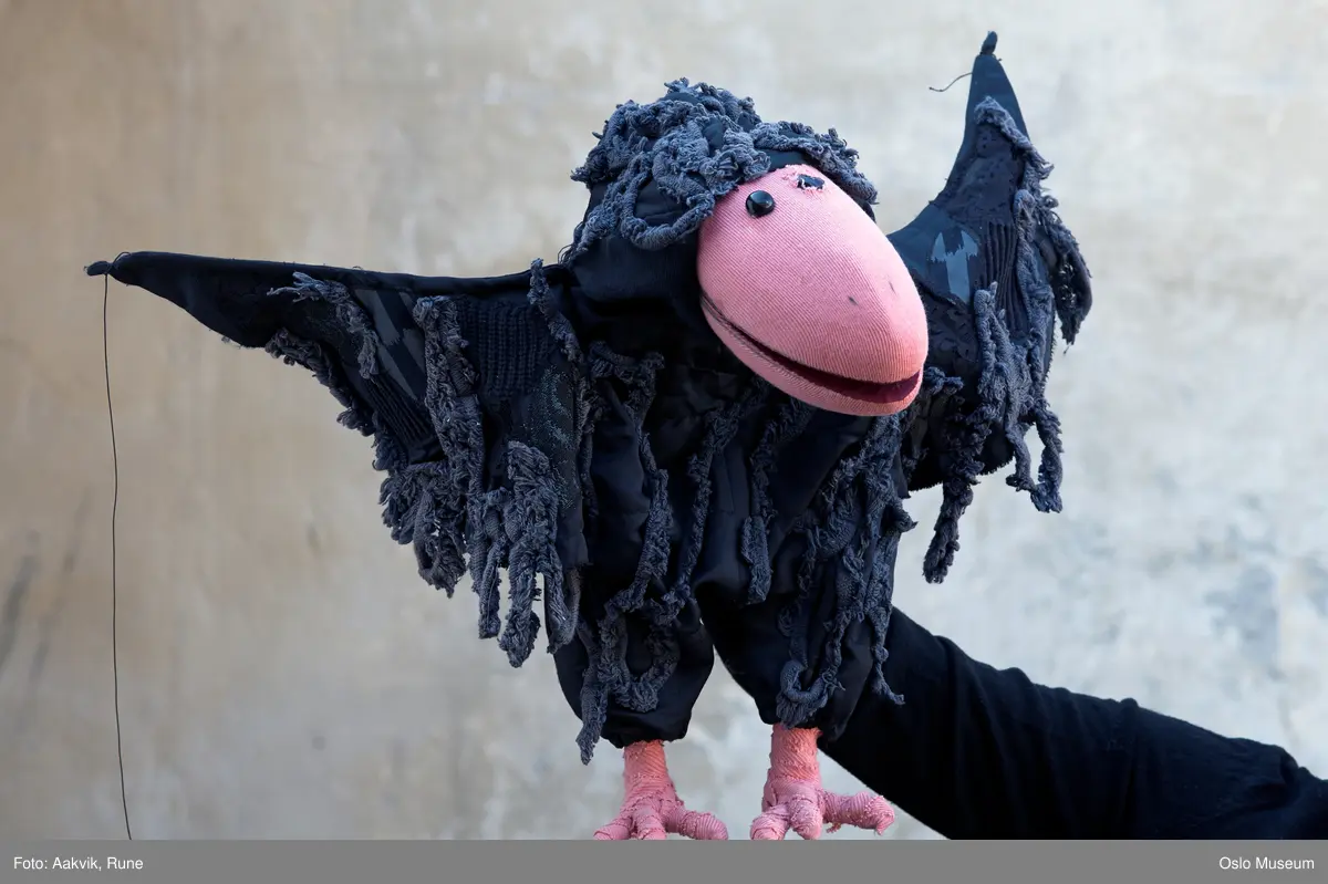 Ravn. Svart med rosa nedd og bein. Hånddukke med hull på ryggen til hånda.
Dukken er noe slitt/ødelagt