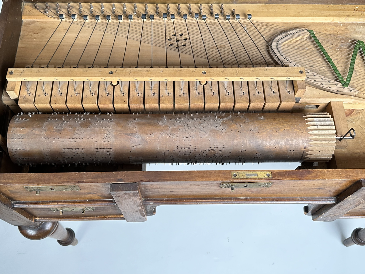 A) Pianoharpa, i form av ett skrivbord av mazarin-typ. Av mörkt betsat och polerat trä. Skrivbordet kraftigt angripet av trägnagare. Svarvade ben och hög sarg, med två utdragbara lådor och tre blindlådor - alla med mässingsbeslag. Bordsskivan uppfällbar och ställbar med metallhake, varunder spelverket med strängar och piggförsedd vals gömmer sig. 
B) och C), två valsar med olika melodier på. Cylinderformade, av trä, med mittaxel och piggar av metall. Valsen snurras med en vev, som lämnat ett svängt märke på möbelns front. 
D), vev av tunn metallarm med ett S-formad avslut i 45 graders vinkel från armen samt svarvad, balusterformat handtag av trä.
E) och F), stämnycklar av metall med fyrkantiga hål, samt enkla handtag av trä. 
G), sprint av metall för att säkra valsen på plats. Tunn, med ögla till avslut.
H), förvaringslåda för melodivals. Av trä, med inbyggd vagga för att hålla valsens mittaxel.