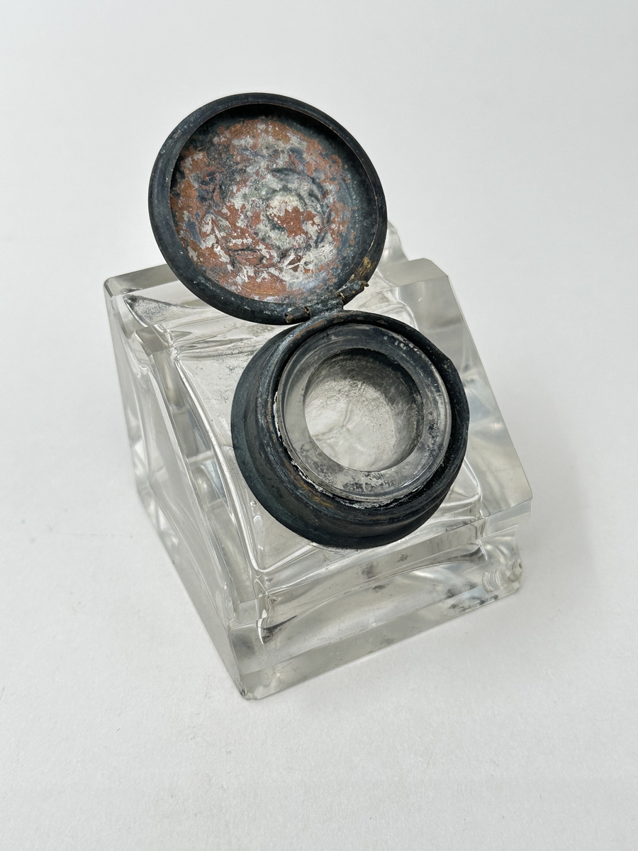 Bläckhorn A med insats B.
Bläckhorn av glas, gjutet i form, med lock av mässing. Modell som en kub med en snett avskuren sida, där locket sitter. Det finns rännor för pennan att vila i på var sida om locket. Locket har ett gångjärn och kan snäppas fast. På ovansidan finns ett pressat motiv, en lagerkrans. Insatsen av glas hänger i locket. Den är trattformad och mäter 31 mm i diameter, 35 mm i höjd. 
Små skador på alla hörn.