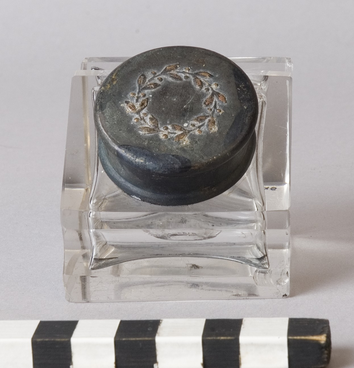 Bläckhorn A med insats B.
Bläckhorn av glas, gjutet i form, med lock av mässing. Modell som en kub med en snett avskuren sida, där locket sitter. Det finns rännor för pennan att vila i på var sida om locket. Locket har ett gångjärn och kan snäppas fast. På ovansidan finns ett pressat motiv, en lagerkrans. Insatsen av glas hänger i locket. Den är trattformad och mäter 31 mm i diameter, 35 mm i höjd. 
Små skador på alla hörn.
