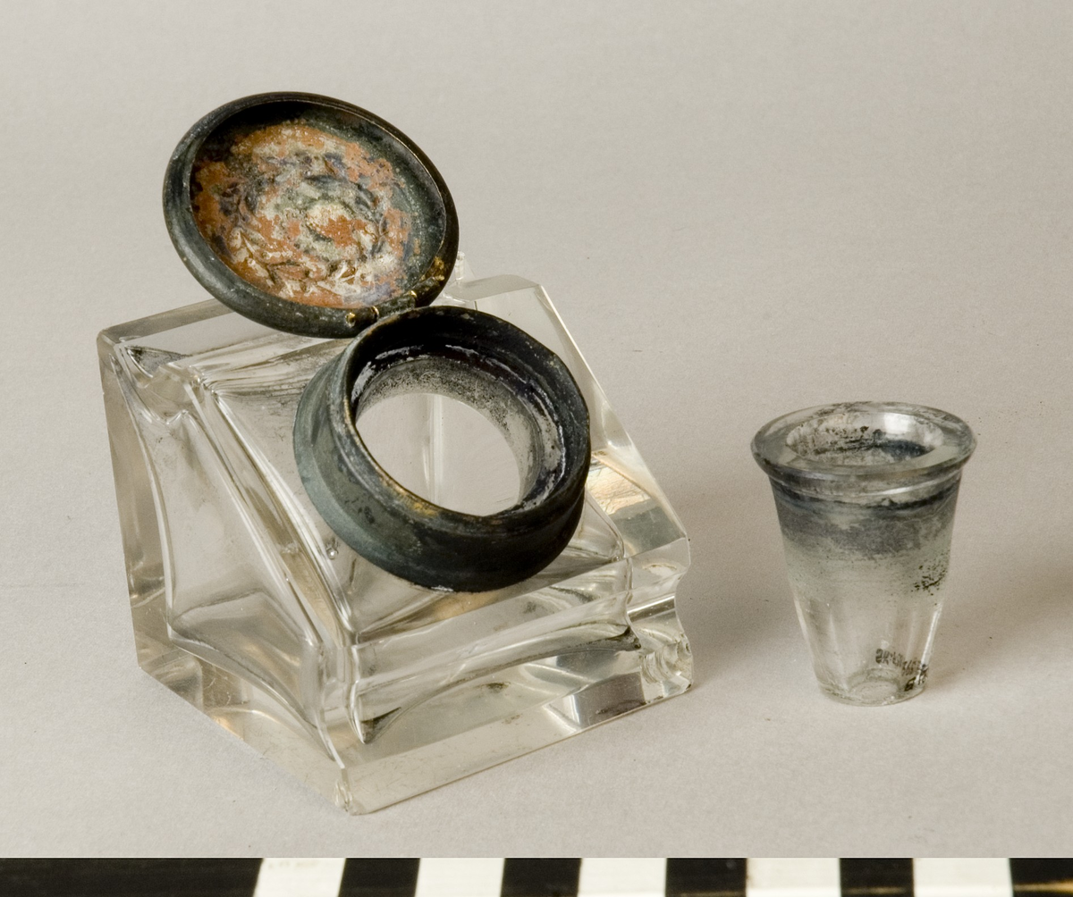 Bläckhorn A med insats B.
Bläckhorn av glas, gjutet i form, med lock av mässing. Modell som en kub med en snett avskuren sida, där locket sitter. Det finns rännor för pennan att vila i på var sida om locket. Locket har ett gångjärn och kan snäppas fast. På ovansidan finns ett pressat motiv, en lagerkrans. Insatsen av glas hänger i locket. Den är trattformad och mäter 31 mm i diameter, 35 mm i höjd. 
Små skador på alla hörn.