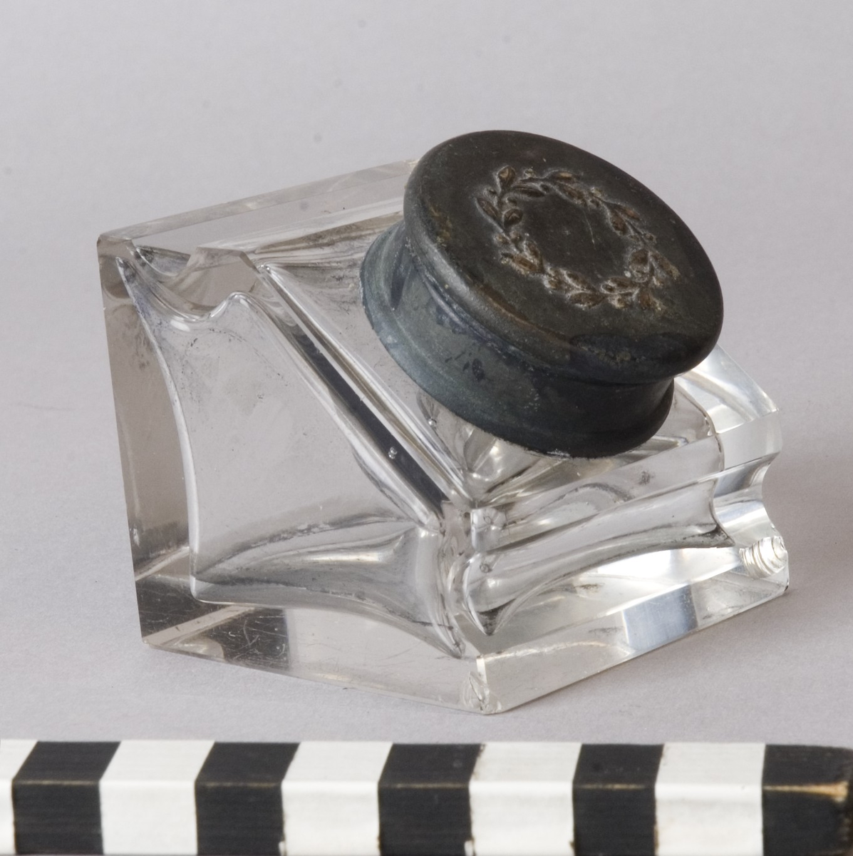 Bläckhorn A med insats B.
Bläckhorn av glas, gjutet i form, med lock av mässing. Modell som en kub med en snett avskuren sida, där locket sitter. Det finns rännor för pennan att vila i på var sida om locket. Locket har ett gångjärn och kan snäppas fast. På ovansidan finns ett pressat motiv, en lagerkrans. Insatsen av glas hänger i locket. Den är trattformad och mäter 31 mm i diameter, 35 mm i höjd. 
Små skador på alla hörn.