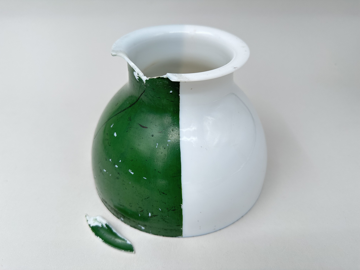 Kupa till rovoljelampa. Av porslin eller möjl. opalglas. Rund med vikt överkant. Till hälften vit och till hälften grönlackerad. Stor A-formad spricka och bit som är borta från kupans övre kant. Skador i lacken.