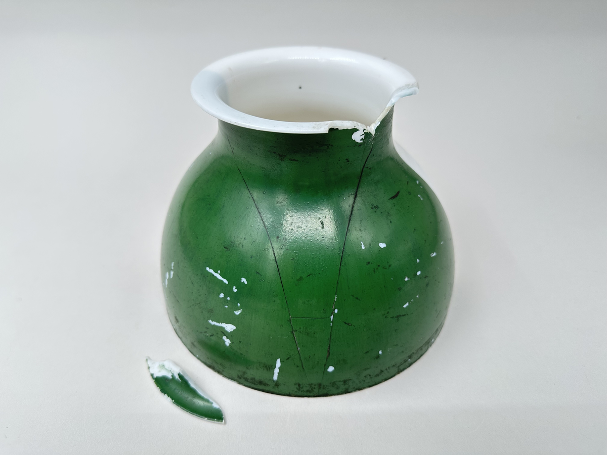 Kupa till rovoljelampa. Av porslin eller möjl. opalglas. Rund med vikt överkant. Till hälften vit och till hälften grönlackerad. Stor A-formad spricka och bit som är borta från kupans övre kant. Skador i lacken.