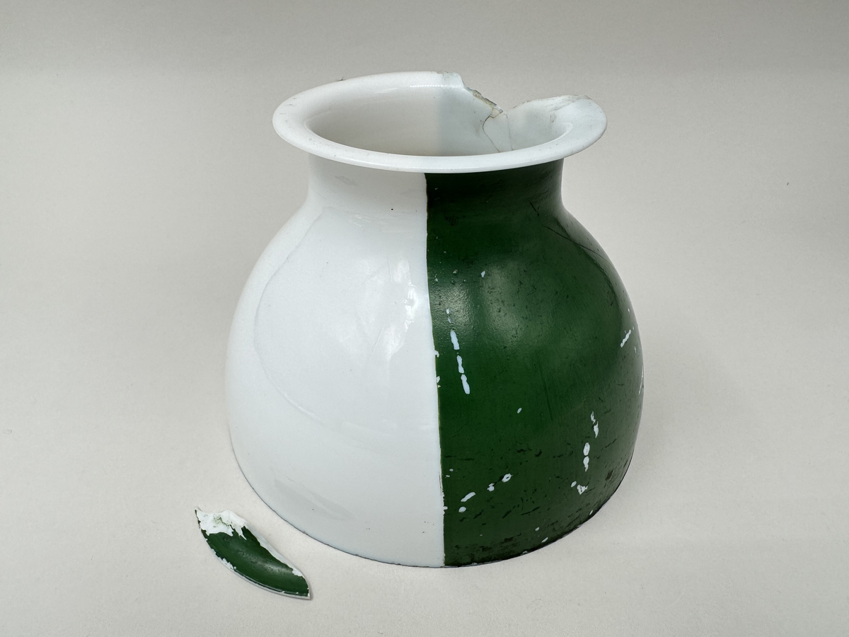 Kupa till rovoljelampa. Av porslin eller möjl. opalglas. Rund med vikt överkant. Till hälften vit och till hälften grönlackerad. Stor A-formad spricka och bit som är borta från kupans övre kant. Skador i lacken.