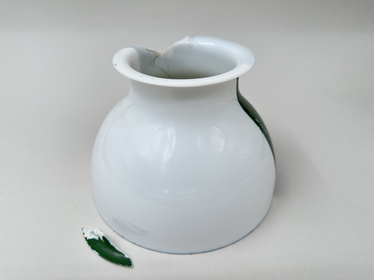 Kupa till rovoljelampa. Av porslin eller möjl. opalglas. Rund med vikt överkant. Till hälften vit och till hälften grönlackerad. Stor A-formad spricka och bit som är borta från kupans övre kant. Skador i lacken.