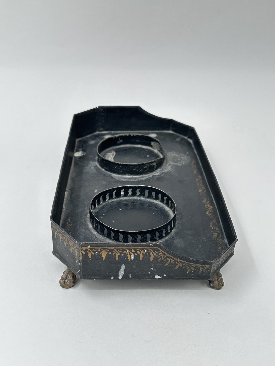 Bricka till skrivtyg, i svartlackerad plåt med målade dekorbårder i guld. Rektangulär med avfasade hörn, ståendes på fyra guldmålade kulklofötter. Två fastlödda rundlar för att hålla bläckhorn och sandströare på plats, den ena rundeln med genombruten dekor.