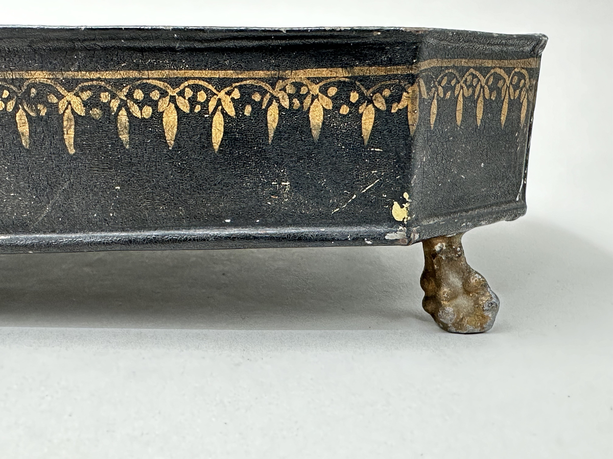 Bricka till skrivtyg, i svartlackerad plåt med målade dekorbårder i guld. Rektangulär med avfasade hörn, ståendes på fyra guldmålade kulklofötter. Två fastlödda rundlar för att hålla bläckhorn och sandströare på plats, den ena rundeln med genombruten dekor.