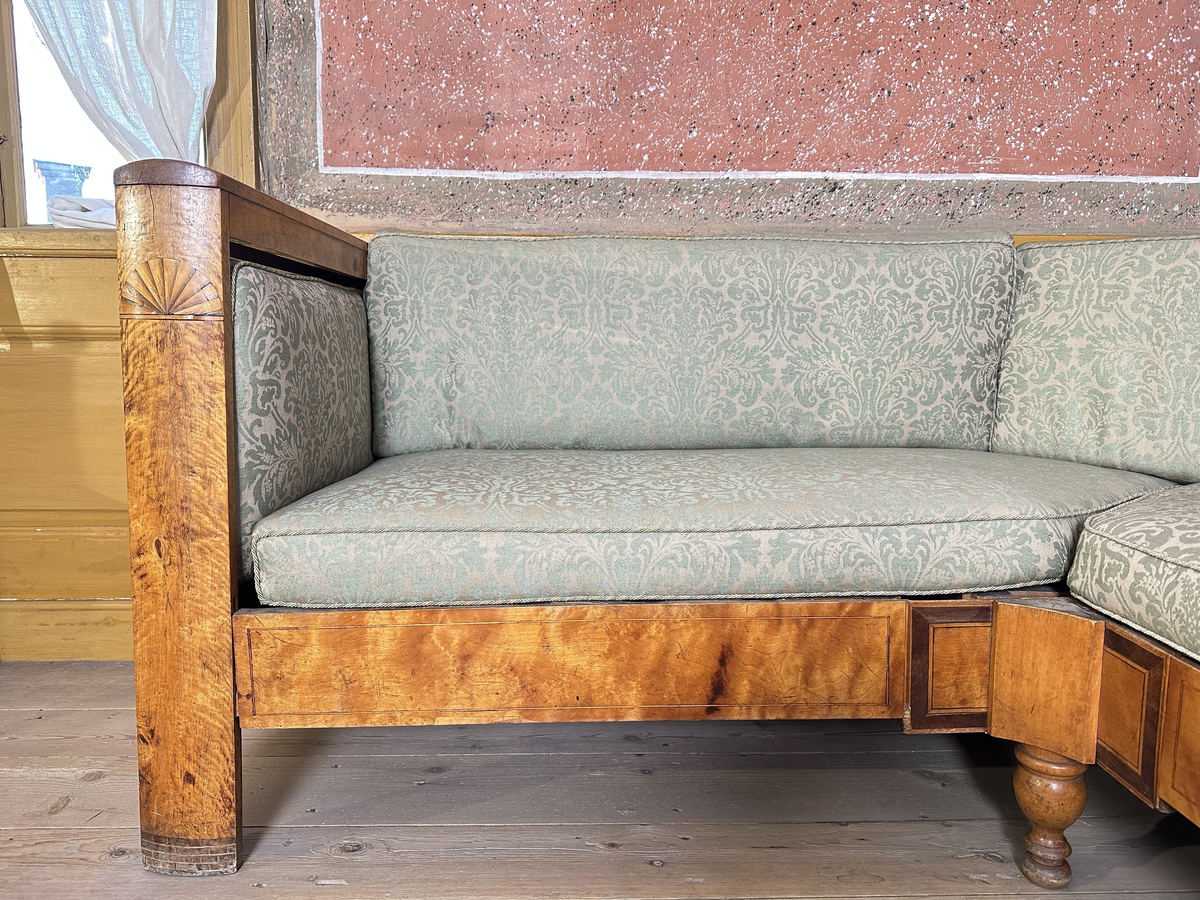 Hörnsoffa i polerad björk med stoppad sits. Genombrutna gavlar med två tvärslåar och spjälor däremellan. Intarsia på gavlarnas kortsidor i form av solfjäderlik halvcirkel samt små rektangulära tegelformade detaljer nederst. Framsargen dekorerad med tunn inläggning i mörkare färg. I hörnets framkant ett svarvat lågt balusterben och något högre sarg med kraftigare inläggning. Ryggen bestående av en sarg baktill med en stödbräda per sida samt en i hörnet. Hörnet klätt i skinnliknande textil och med upphöjd kvadrat i mörkt trä (senare tillkommen). Ribbotten. Sitsar och ryggdynor klädda i lätt glansigt tyg med blomsterornamentik i grön och beige. Kantsnodd i samma kulörer. Baksida i juteväv. 