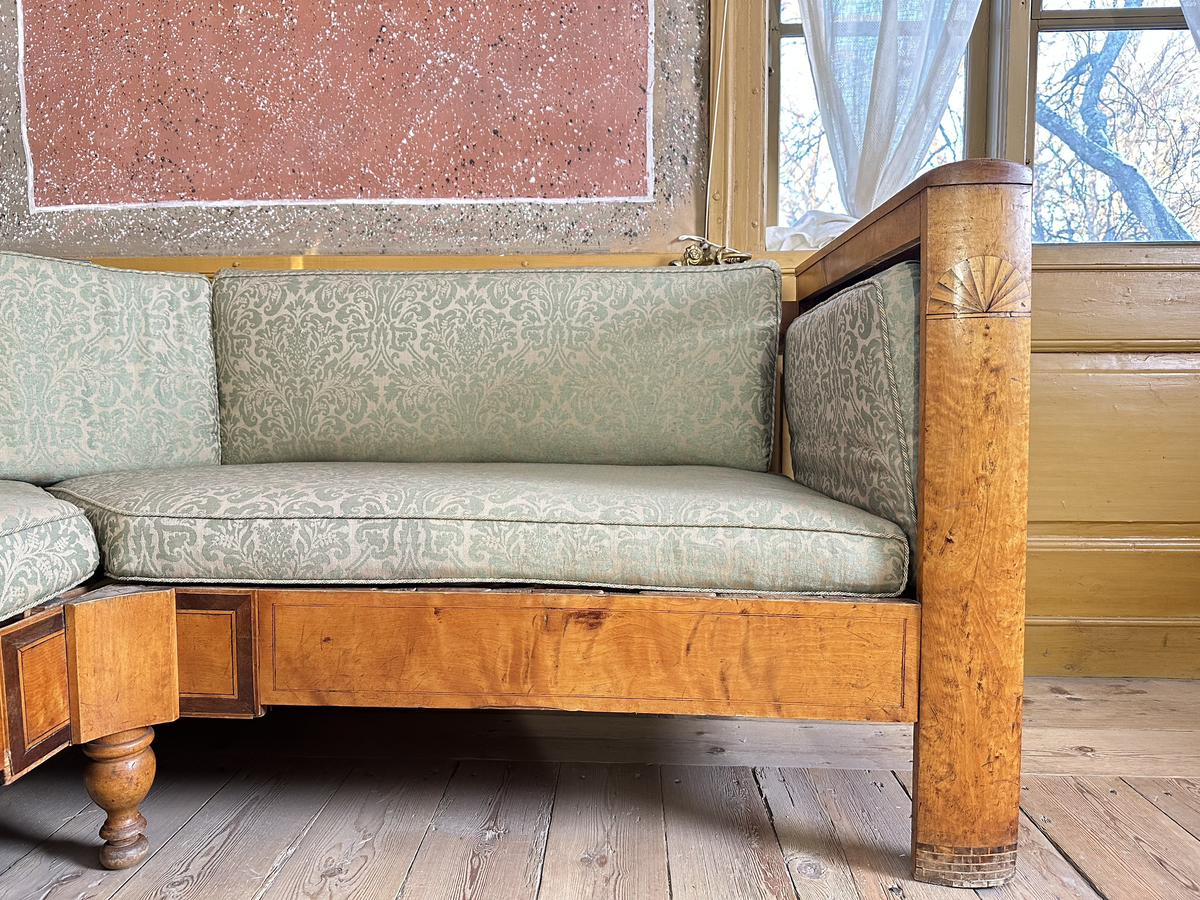 Hörnsoffa i polerad björk med stoppad sits. Genombrutna gavlar med två tvärslåar och spjälor däremellan. Intarsia på gavlarnas kortsidor i form av solfjäderlik halvcirkel samt små rektangulära tegelformade detaljer nederst. Framsargen dekorerad med tunn inläggning i mörkare färg. I hörnets framkant ett svarvat lågt balusterben och något högre sarg med kraftigare inläggning. Ryggen bestående av en sarg baktill med en stödbräda per sida samt en i hörnet. Hörnet klätt i skinnliknande textil och med upphöjd kvadrat i mörkt trä (senare tillkommen). Ribbotten. Sitsar och ryggdynor klädda i lätt glansigt tyg med blomsterornamentik i grön och beige. Kantsnodd i samma kulörer. Baksida i juteväv. 