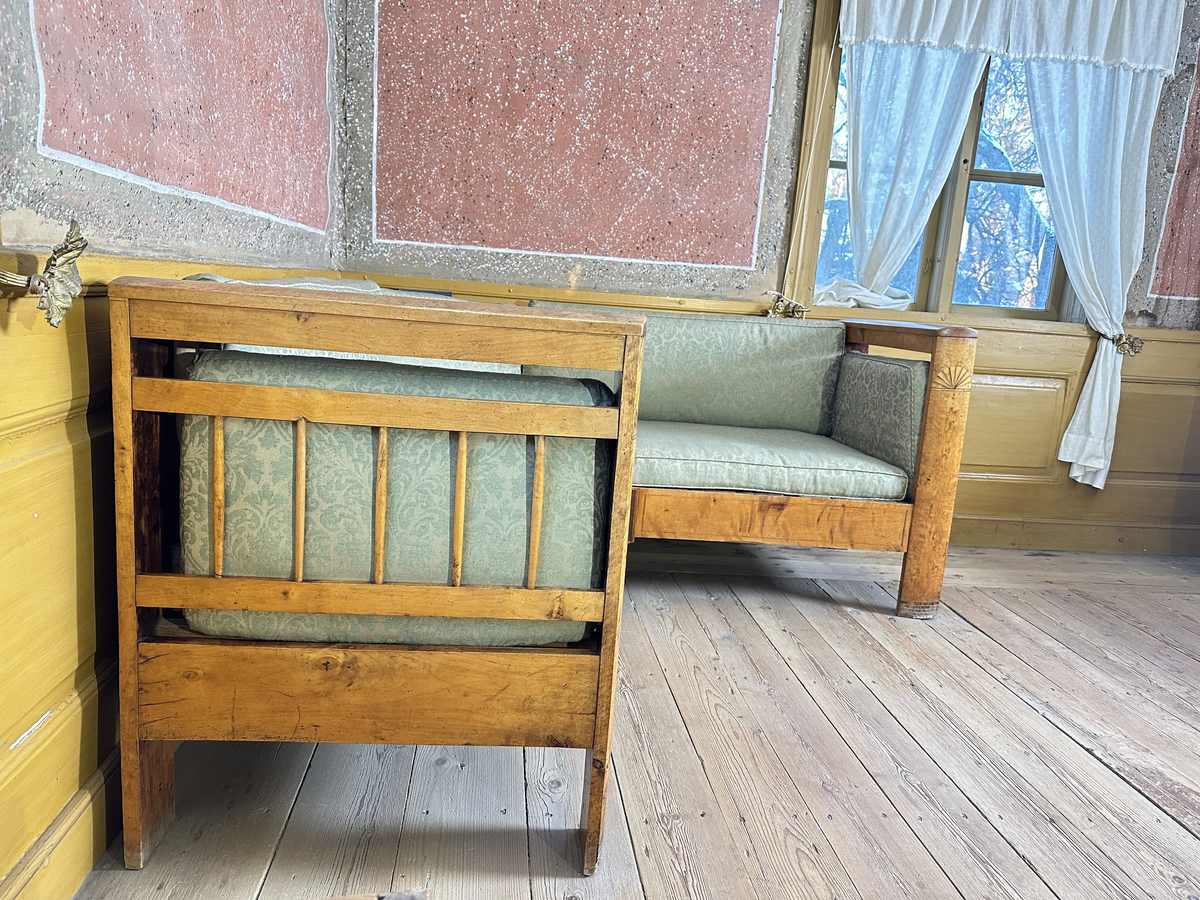 Hörnsoffa i polerad björk med stoppad sits. Genombrutna gavlar med två tvärslåar och spjälor däremellan. Intarsia på gavlarnas kortsidor i form av solfjäderlik halvcirkel samt små rektangulära tegelformade detaljer nederst. Framsargen dekorerad med tunn inläggning i mörkare färg. I hörnets framkant ett svarvat lågt balusterben och något högre sarg med kraftigare inläggning. Ryggen bestående av en sarg baktill med en stödbräda per sida samt en i hörnet. Hörnet klätt i skinnliknande textil och med upphöjd kvadrat i mörkt trä (senare tillkommen). Ribbotten. Sitsar och ryggdynor klädda i lätt glansigt tyg med blomsterornamentik i grön och beige. Kantsnodd i samma kulörer. Baksida i juteväv. 