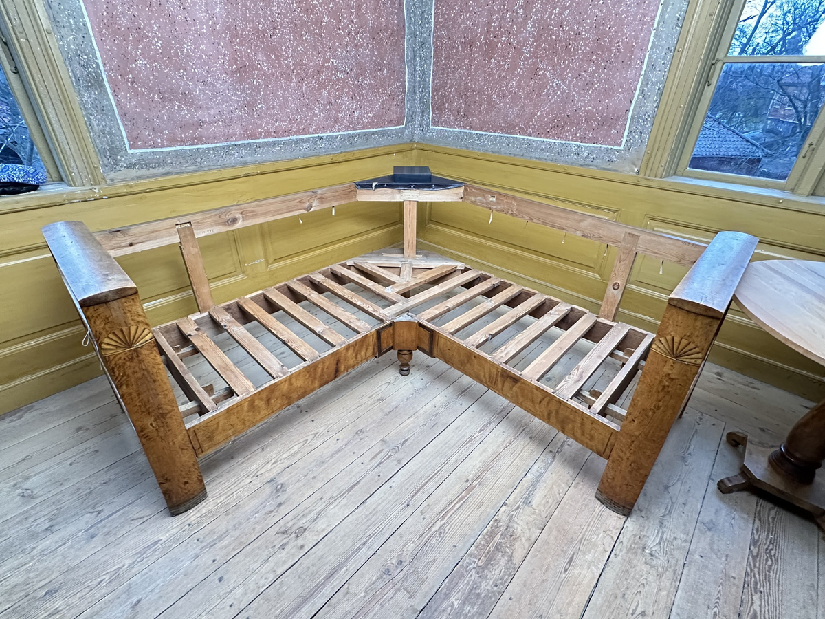 Hörnsoffa i polerad björk med stoppad sits. Genombrutna gavlar med två tvärslåar och spjälor däremellan. Intarsia på gavlarnas kortsidor i form av solfjäderlik halvcirkel samt små rektangulära tegelformade detaljer nederst. Framsargen dekorerad med tunn inläggning i mörkare färg. I hörnets framkant ett svarvat lågt balusterben och något högre sarg med kraftigare inläggning. Ryggen bestående av en sarg baktill med en stödbräda per sida samt en i hörnet. Hörnet klätt i skinnliknande textil och med upphöjd kvadrat i mörkt trä (senare tillkommen). Ribbotten. Sitsar och ryggdynor klädda i lätt glansigt tyg med blomsterornamentik i grön och beige. Kantsnodd i samma kulörer. Baksida i juteväv. 