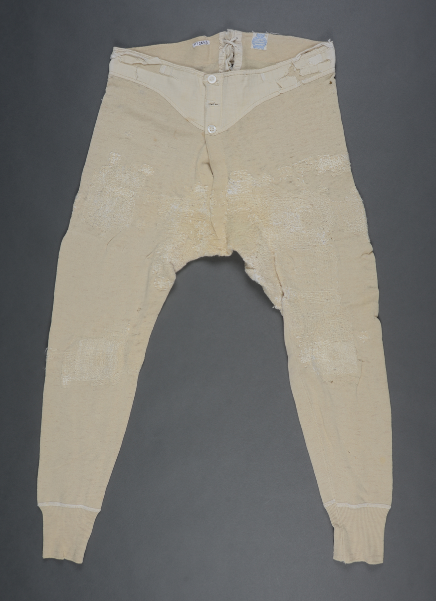 Langunderbukse i naturfarget ull. Påsydd forsterkning av bomull i linningen. Gylf med tre knapper. Splitt bak med snøring. Plagget bærer preg av mye bruk i form av slitasje og mye lapping/stopping foran og bak med ull- og bomullsgarn.
