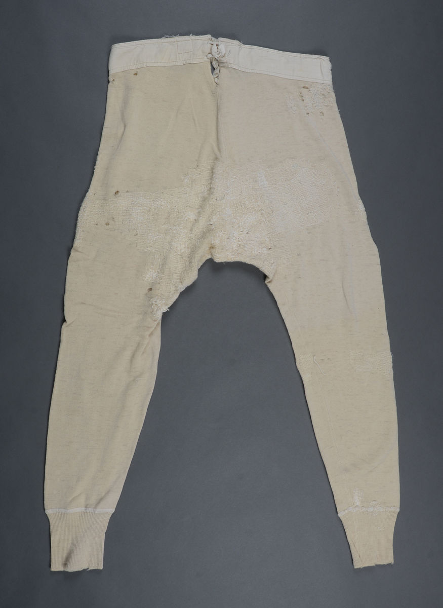 Langunderbukse i naturfarget ull. Påsydd forsterkning av bomull i linningen. Gylf med tre knapper. Splitt bak med snøring. Plagget bærer preg av mye bruk i form av slitasje og mye lapping/stopping foran og bak med ull- og bomullsgarn.