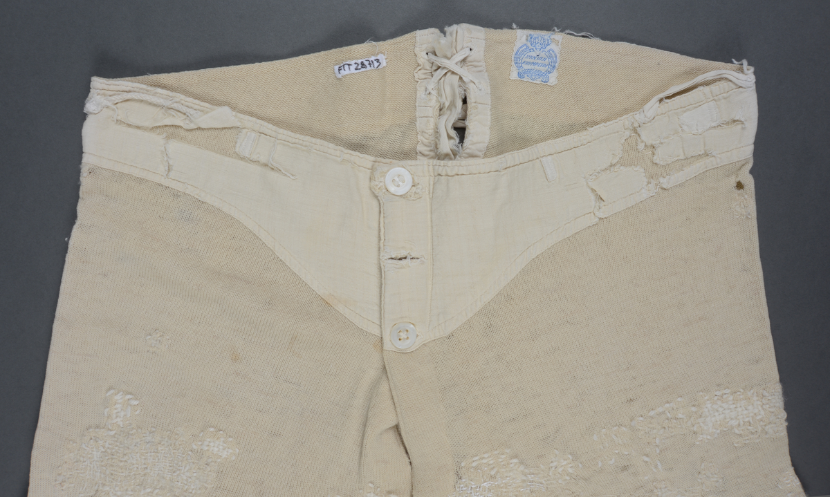 Langunderbukse i naturfarget ull. Påsydd forsterkning av bomull i linningen. Gylf med tre knapper. Splitt bak med snøring. Plagget bærer preg av mye bruk i form av slitasje og mye lapping/stopping foran og bak med ull- og bomullsgarn.