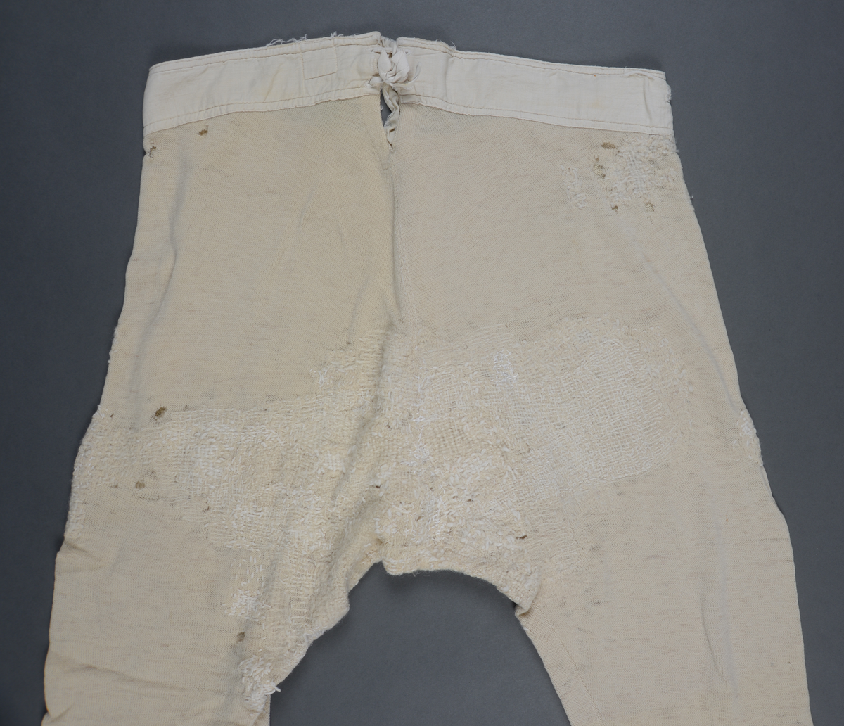 Langunderbukse i naturfarget ull. Påsydd forsterkning av bomull i linningen. Gylf med tre knapper. Splitt bak med snøring. Plagget bærer preg av mye bruk i form av slitasje og mye lapping/stopping foran og bak med ull- og bomullsgarn.