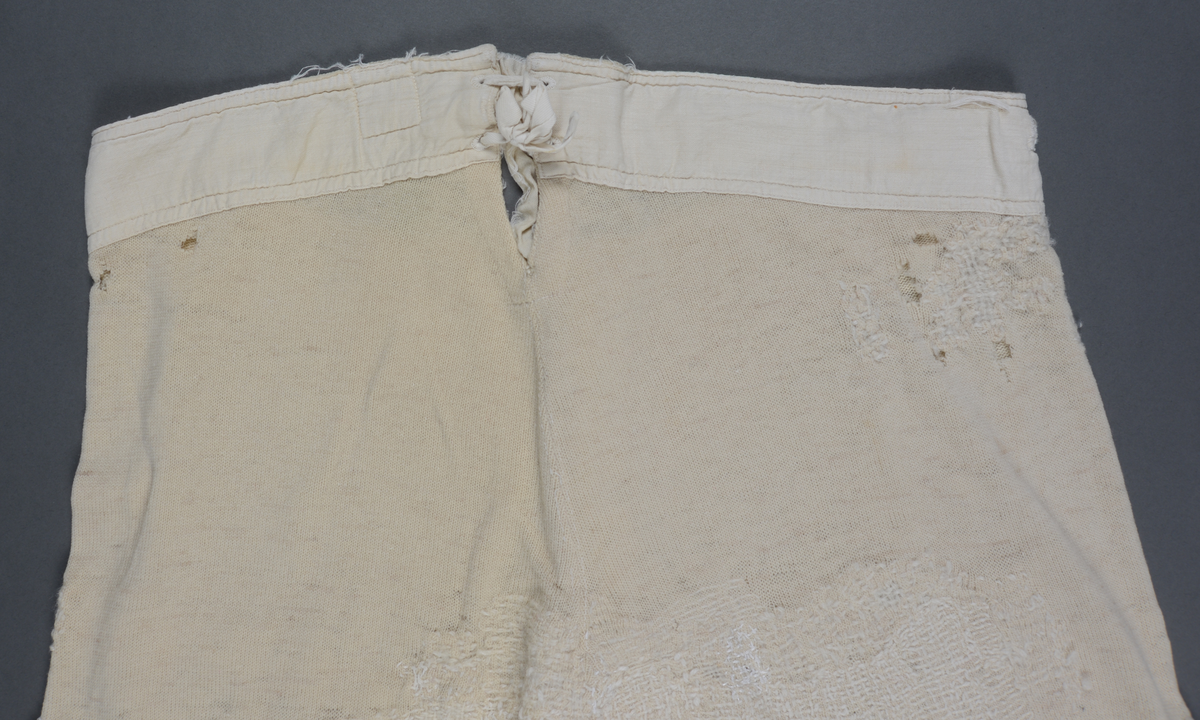 Langunderbukse i naturfarget ull. Påsydd forsterkning av bomull i linningen. Gylf med tre knapper. Splitt bak med snøring. Plagget bærer preg av mye bruk i form av slitasje og mye lapping/stopping foran og bak med ull- og bomullsgarn.