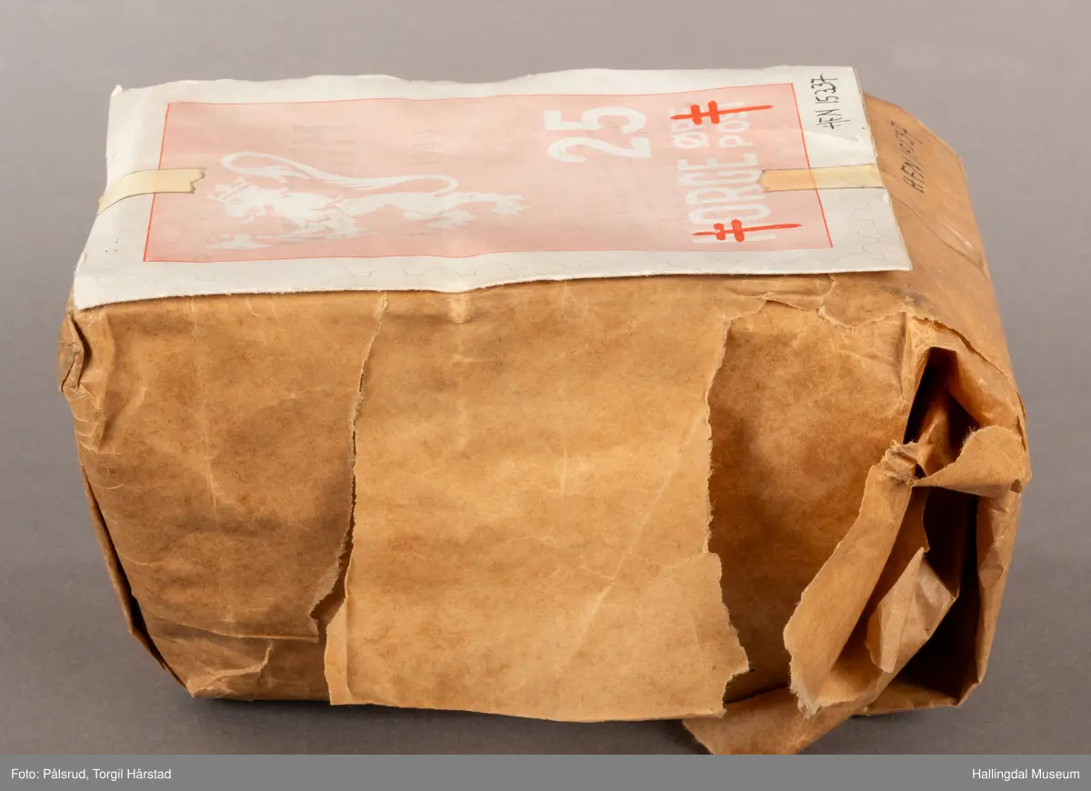 En pakke med uåpne trykksaker til Tubfrim, blant annet årsberetningen for 1949. Pakken er pakket inn med kraftpapir med illustrasjonen til forsiden til årsberetningen utenpå: Et frimerkemotiv med en hvit riksløve på lyserød bakgrunn, under står det Norge og 25 øre post. I tillegg er det plassert to røde Lothringenkors, eller dobbeltkors, i de nederste hjørnene.