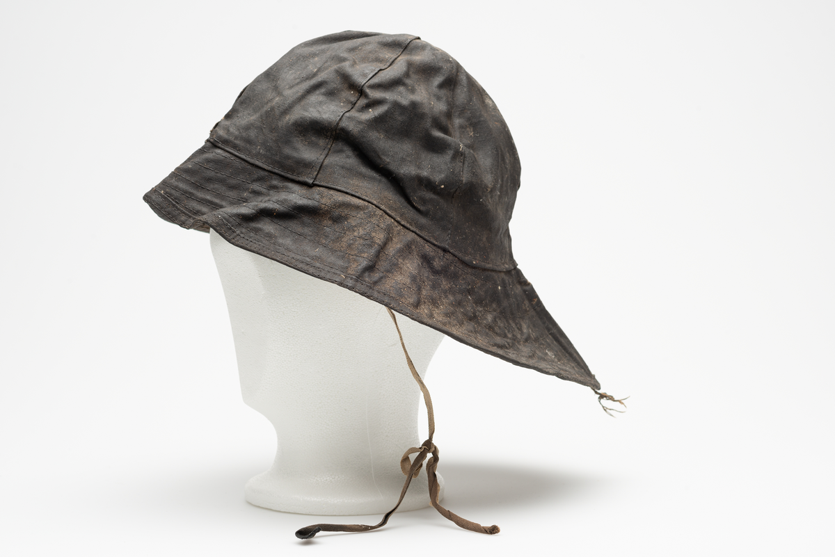 Sydvest i svart oljelerret(oilskin), linoljebehandlet bomullslerret. En avstivet brem, bredere bak enn foran. Foret med hvit/beige bomullsflanel. Øreflapper med knytebånd. Hakesnor reparert av bruker. Opphengshempe midt bak på brem.