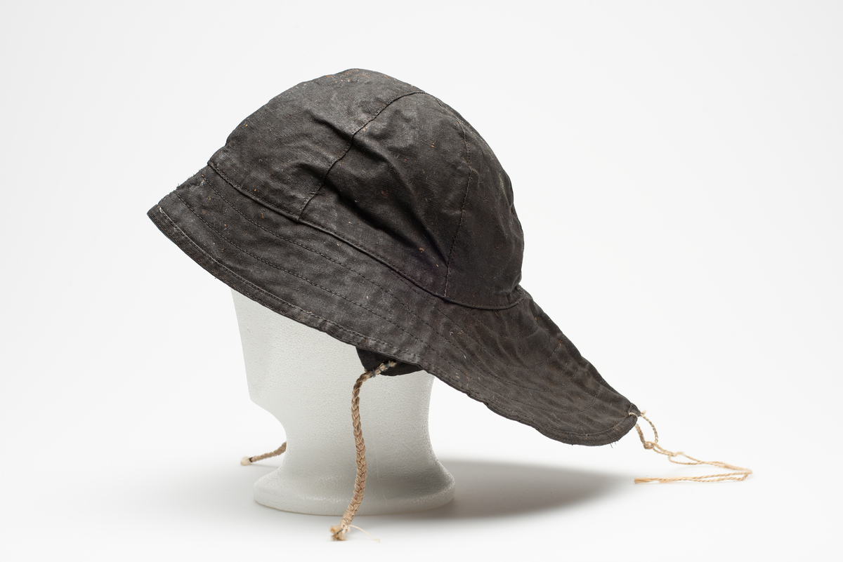 Svart sydvest. Avstivet brem, bredere bak en foran. Invendig foret med naturfarget bomullsflanell. Innvendige øreflapper med hvitt flettet knytebånd. Flettet hvit opphengshempe midt bak på brem. Svak logo fra Helly Hansen på venstre øreflapp.