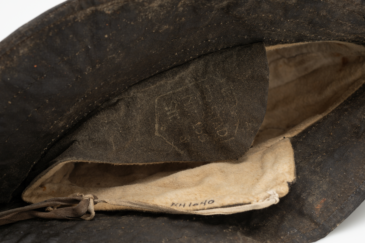 Sydvest i svart oljelerret(oilskin), linoljebehandlet bomullslerret. En avstivet brem, bredere bak enn foran. Foret med hvit/beige bomullsflanel. Øreflapper med knytebånd. Hakesnor reparert av bruker. Opphengshempe midt bak på brem.