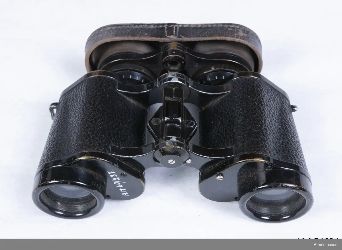 Dubbelkikare för militärt bruk av märket Carl Zeiss Jena Nedinsco samt tillhörande fodral. Kikarfodralet är av märket Busch och samhör inte ursprungligen med själva kikaren. 

´s-Gravenhage är det äldre officiella namnet på Haag, vilket står inpräntat på föremålet.

Märkningen ”A” står högst troligt för Artilleriet. Kronmärkt vilket stämplades redan vid tillverkningen i fabriken i Nederländerna. Övriga kikarmodeller i serien beställdes av svenska flottan i 7x50 förstoring och för danska försvaret i 8x40.
6x30: Siffran 6 avser själva förstoringen alternativt att det iakttagna föremålet i kikaren upplevs vara 6 gånger närmare än i verkligheten. 30 avser diametern i millimeter på frontlinsen längst fram på kikaren där ljusinsläppet sker. 

Exemplaret har extra stora ögonlinser vilket underlättar hanteringen då kikaren ska användas i kombination med gasmask. Kikaren kan då inte läggas an direkt mot ögonen utan måste hållas mot gasmasken. Föremålet är det enda kända kvarvarande i den här förstoringen i kombination med den särskilda bygeln på ovansidan avsedd att manövrera gasmaskokularet fram och tillbaka. Konstruktionen med bygeln fungerade inte särskilt väl eftersom den både var skör och svårmanövrerad, varpå bygeln senare togs bort. Exemplaret skiljer sig gentemot andra liknande kikarmodeller som har vita gasmaskokular. 

Vid observation i kikaren kan den särskilda streckskalan skönjas i det vänstra okularet med numrering 0-100 horisontellt och 0-20 vertikalt. Hjälpmedlet underlättar avståndsbedömningar av verkliga föremål i terrängen eller över vattenytan. 

Grupp F III.
Kikare märkt "Systeem Nedinsco Carl Zeiss Jena S. Gravenhage 1474936" och "Krona A:2084 6x30".